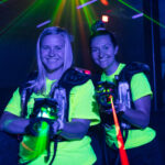 Two woman in neon yellow shirts wearing laser tag equipment with their phasers facing the camera, one with a green auora laser and one with the red aiming laser.
