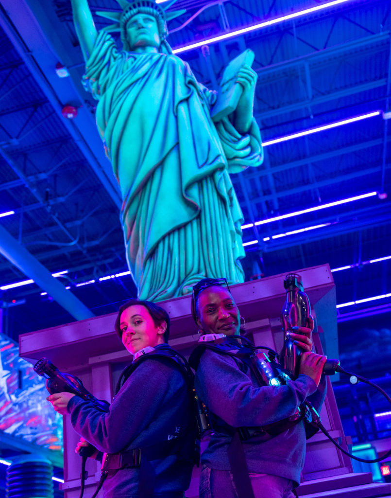 Two women wearing laser tag equipment standing in front of a statue of liberty statue.