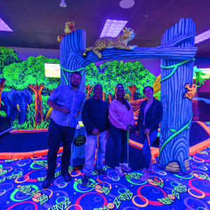 Four people standing in a row under a decorative archway on a glow-in-the-dark mini golf course.