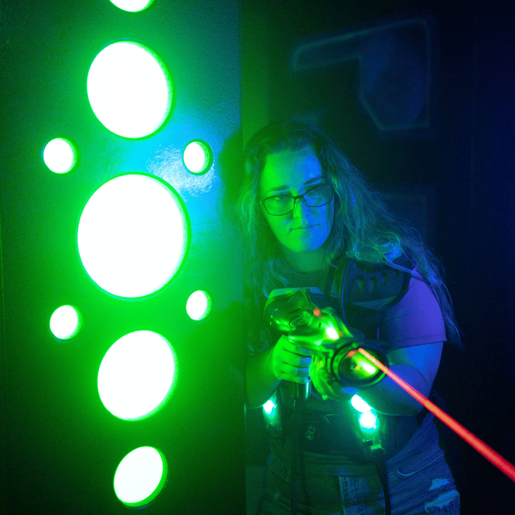 A woman in CyberBlast Pro laser tag equipment firing a red aiming laser from behind a green glowing wall.