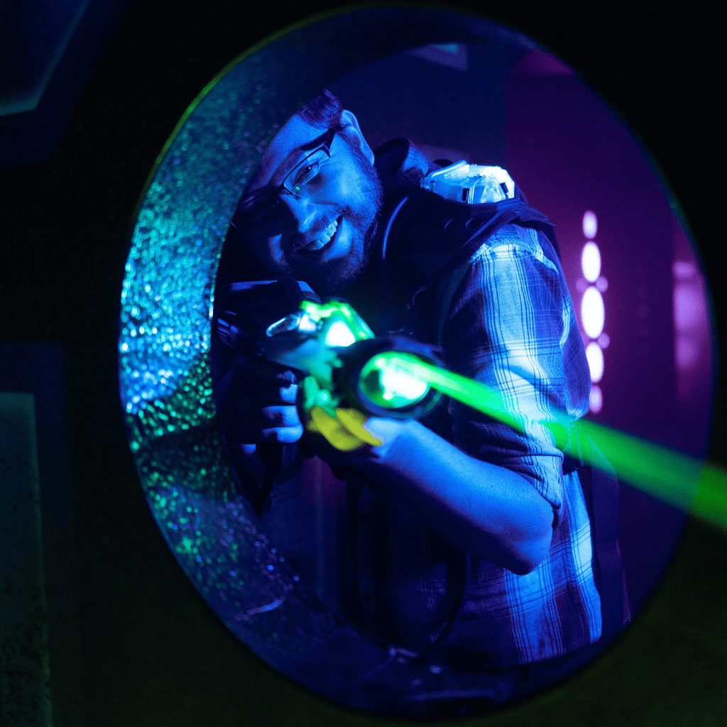 Man in laser tag gear smiling and shooting a green laser through a wall cutout.