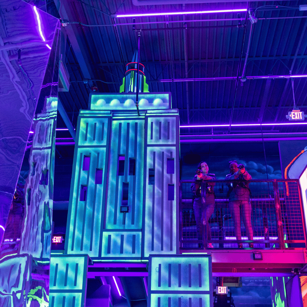 Laser tag arena with a large building prop and a bridge connected to it with two woman pointing laser tag equipment down at the camera.