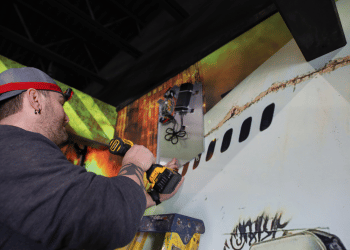 Man on a ladder using a hand drill to install a laser tag base.