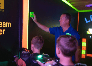 Man pushing a start button on the wall of a laser tag vesting room while two children walk wearing laser tag equipment walk into the arena.