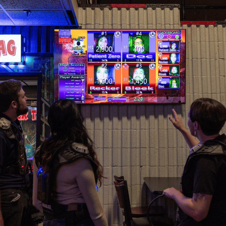 Three people standing with their backs facing the camera as they look and point at the photos on a laser tag scoreboard.